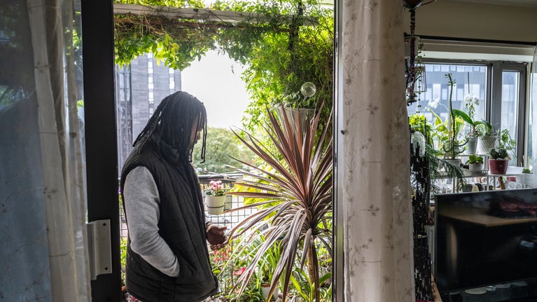 One of the judges of the National Trust's Sky Gardening Challenge 2025 stands in the doorway to a balcony which is covered in lush green plants. Beyond is a view of the Manchester skyline.